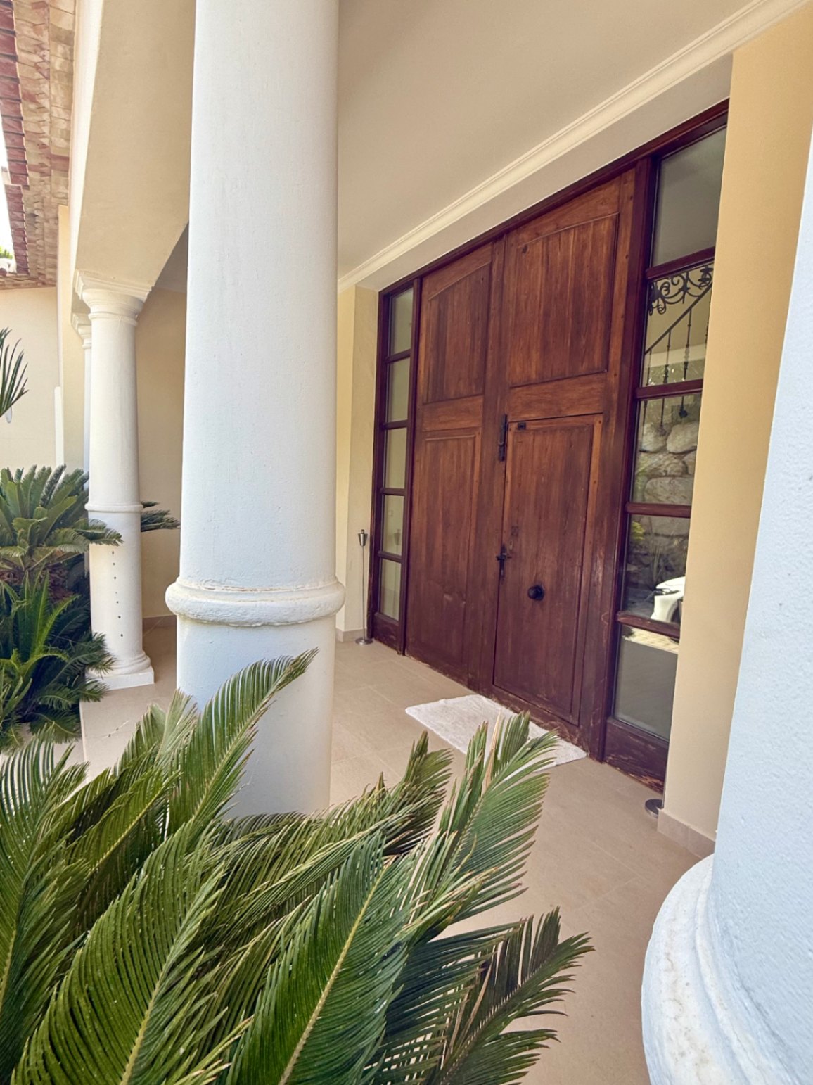 Entrada principal con imponente puerta de madera maciza, flanqueada por ventanales verticales y columnas blancas de estilo toscano entre vegetación mediterránea.