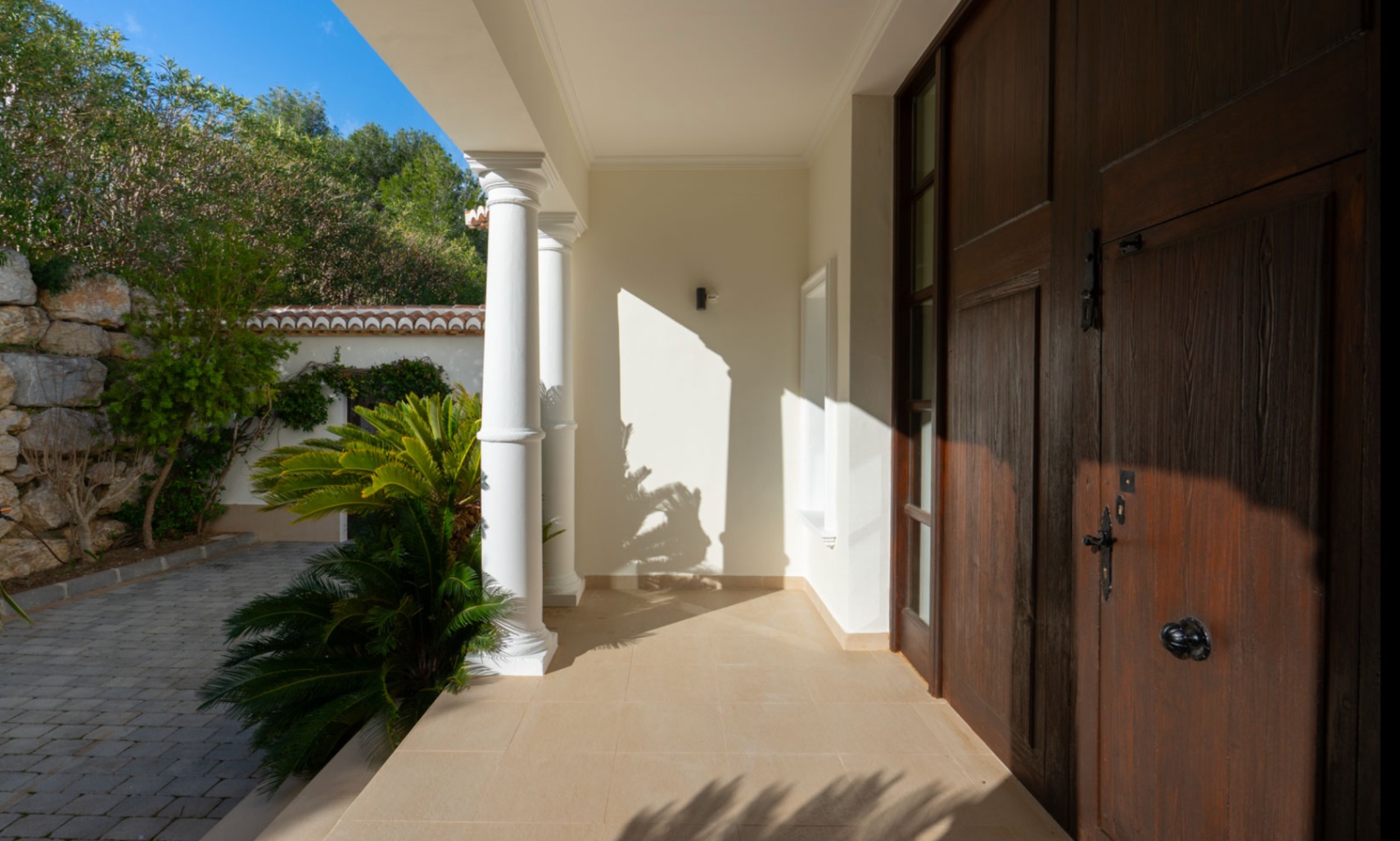 Entrada principal con puerta de madera maciza y columnas blancas clásicas. El porche cubierto destaca por su suelo cerámico y vistas al acceso empedrado con vegetación.