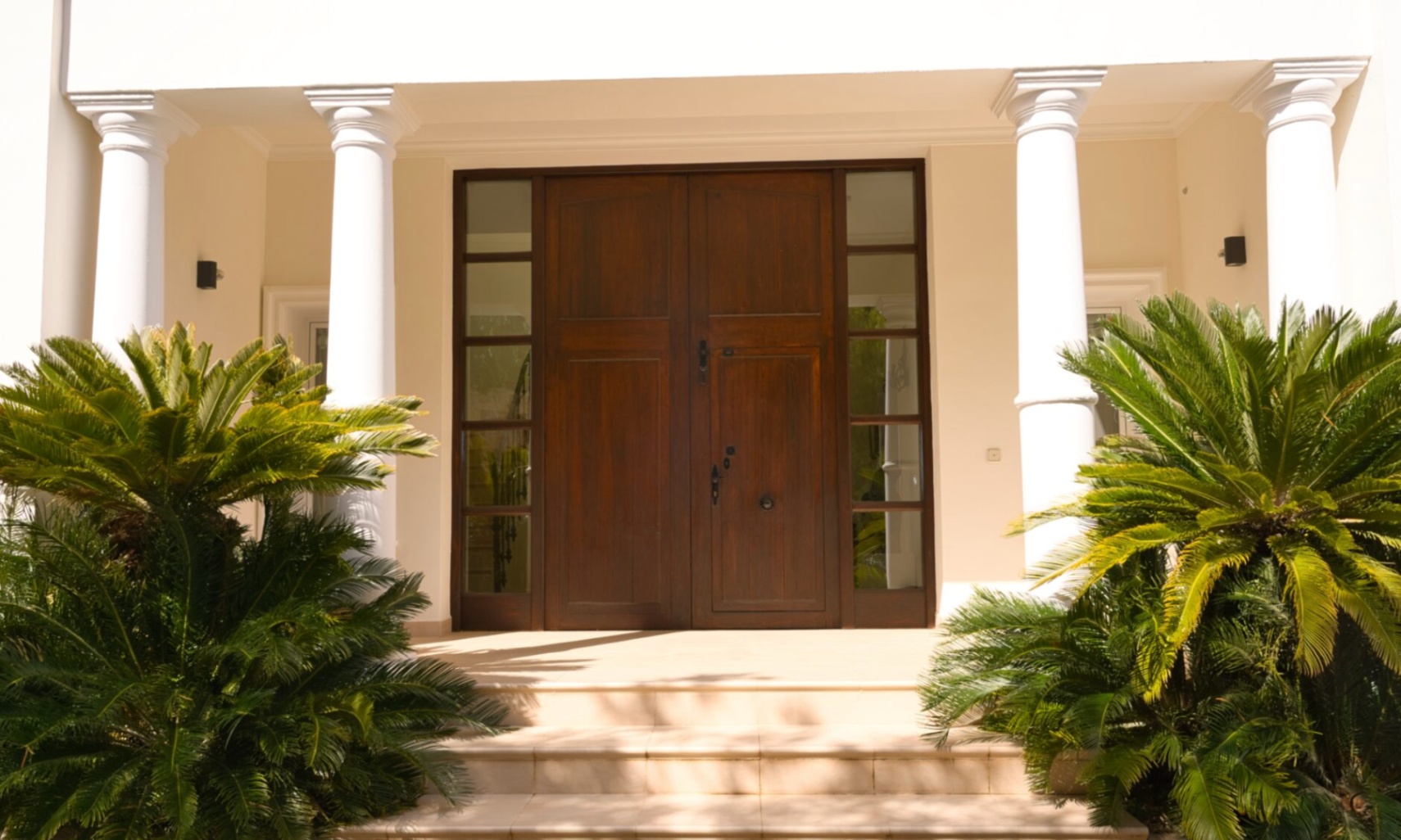 Entrada principal con puerta doble de madera oscura flanqueada por ventanales laterales, columnas blancas de estilo clásico y vegetación mediterránea sobre escalones de piedra.