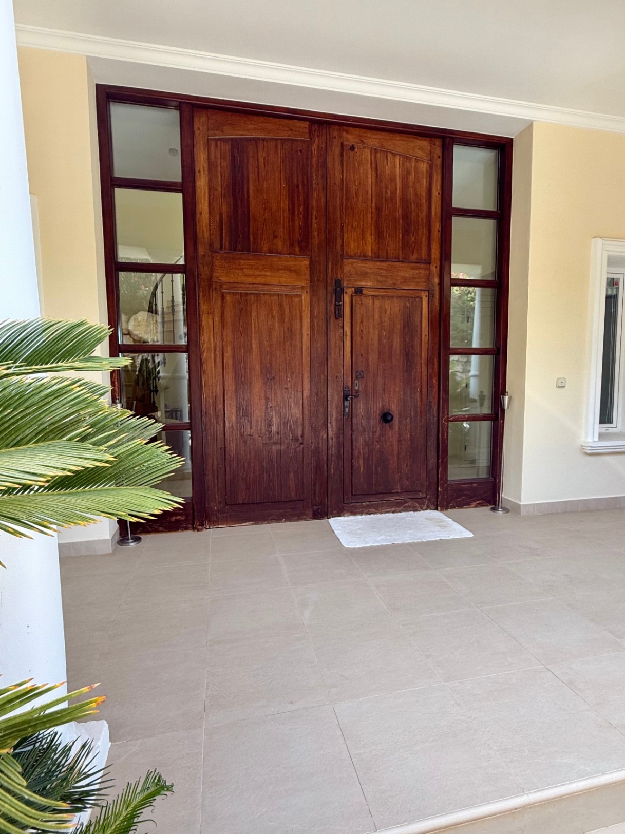 Entrada principal con una gran puerta de madera maciza oscura, flanqueada por ventanales laterales y un porche de baldosas cerámicas claras.