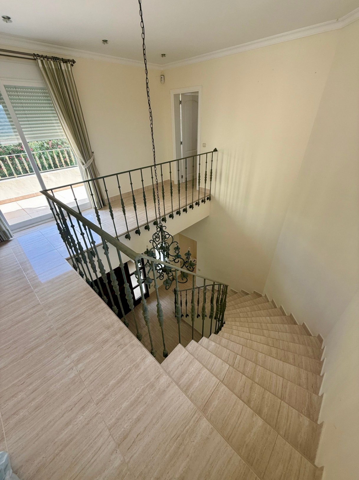 Escalera curva de mármol con barandilla de hierro forjado y una lámpara de araña central, conectando con un rellano luminoso con acceso a la terraza.
