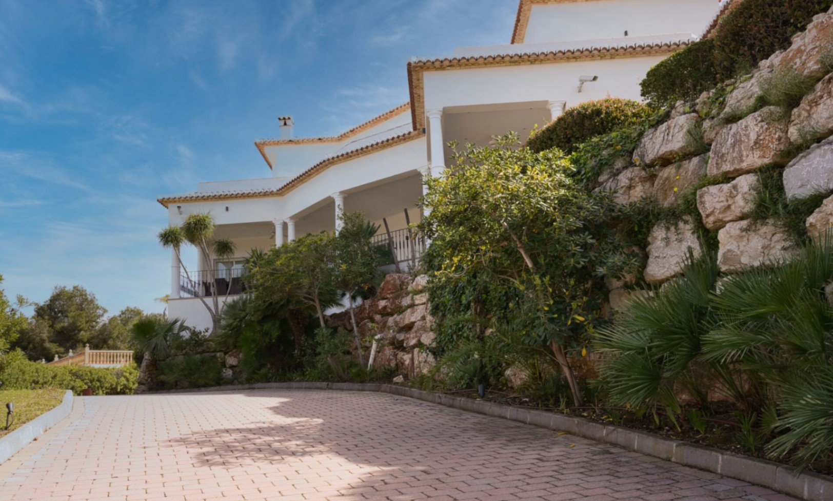 Fachada mediterránea con amplias terrazas cubiertas y columnas, flanqueada por un muro de piedra natural y acceso pavimentado rodeado de vegetación autóctona.