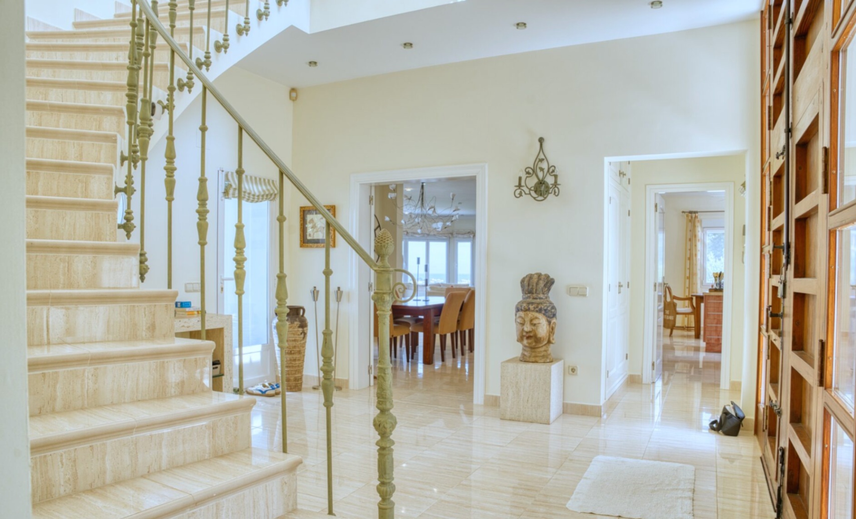 Hall de entrada con escalera de mármol y barandilla de hierro forjado. Suelos de mármol pulido conectan con el comedor y ofrecen vistas al mar al fondo.