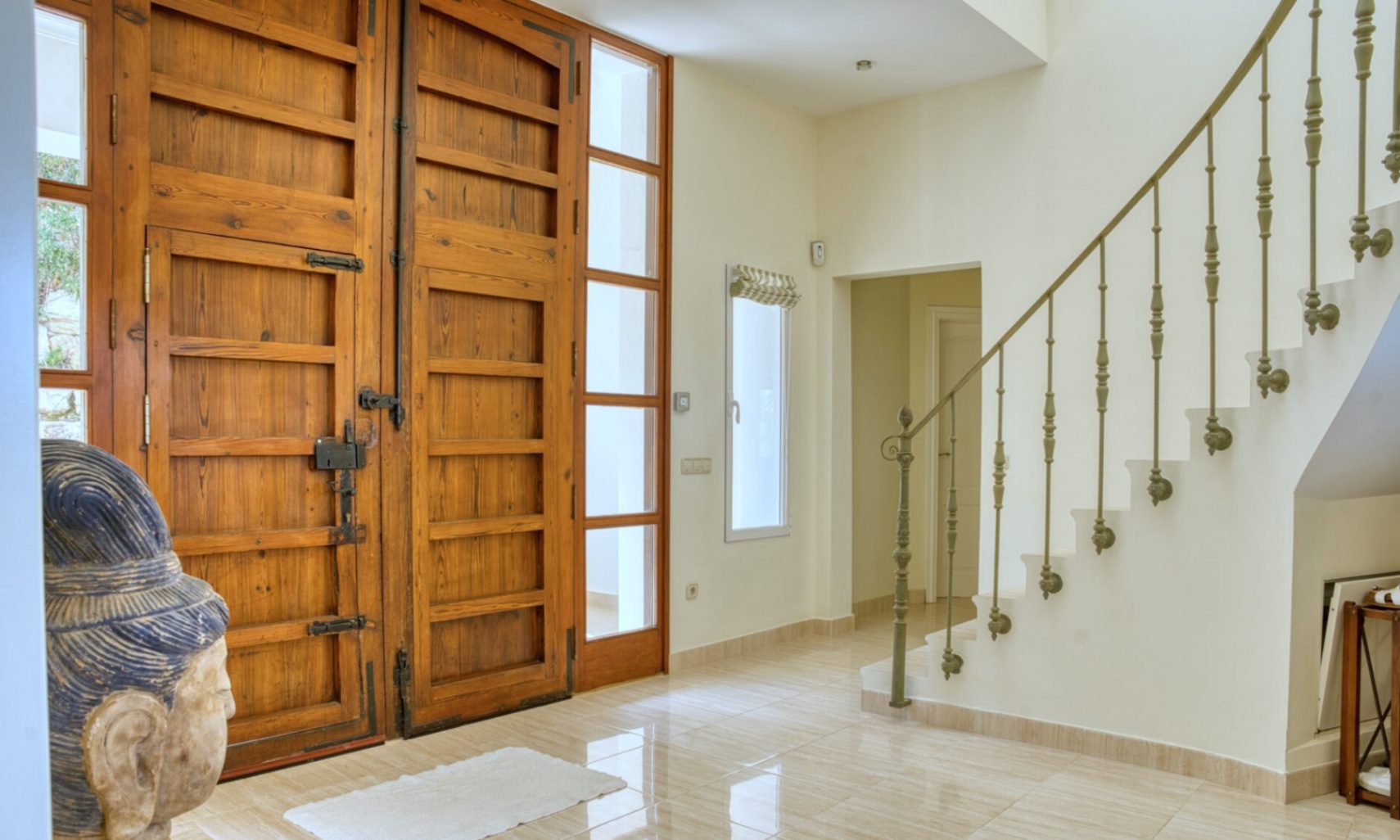 Hall de entrada con puertas dobles de madera rústica, herrajes de hierro forjado y suelos de gres pulido. Escalera con barandilla ornamental y abundante luz natural.