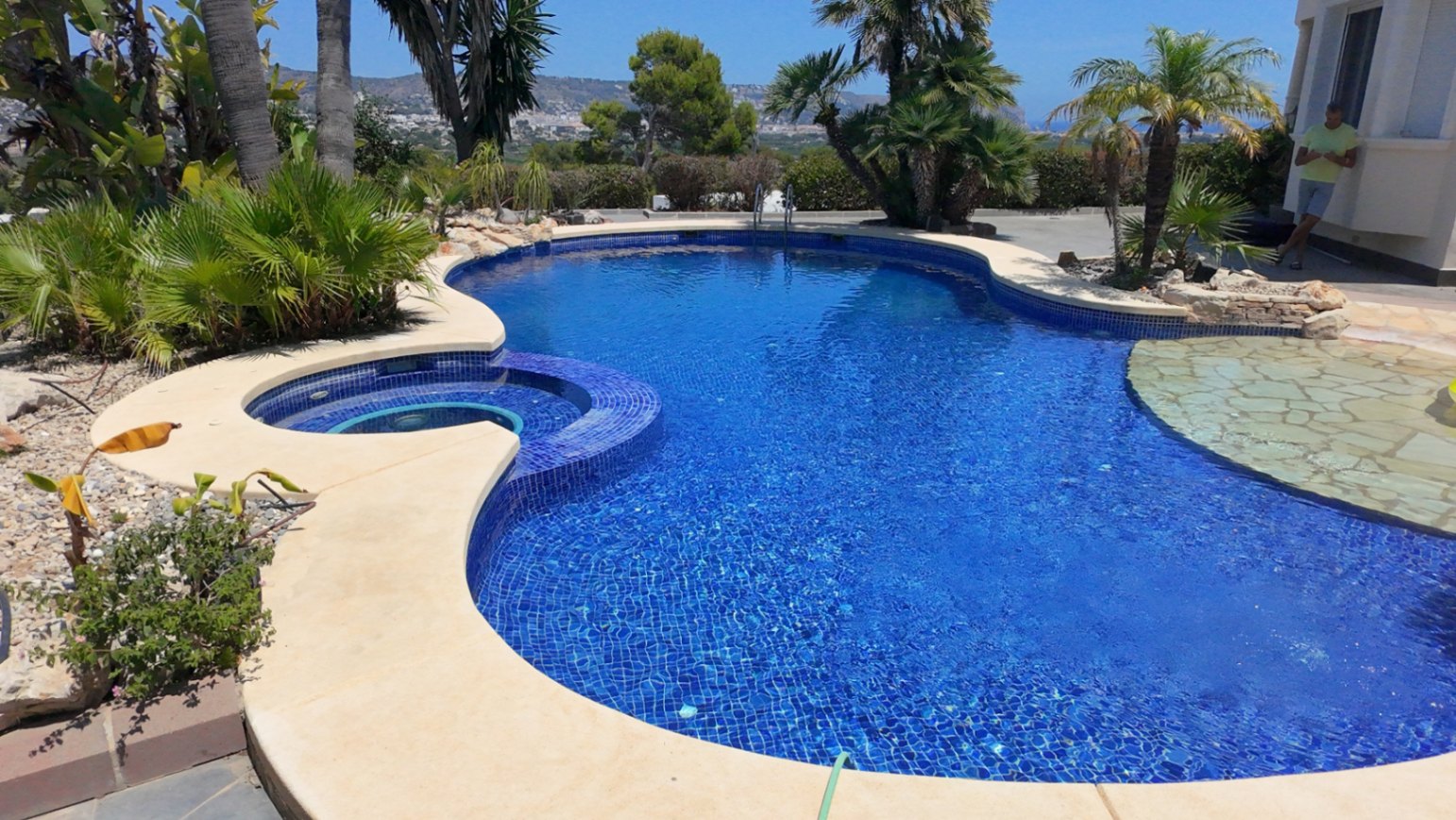 Piscina de forma orgánica con mosaico azul, jacuzzi circular integrado y zona de entrada en piedra natural, rodeada de palmeras y vegetación mediterránea.