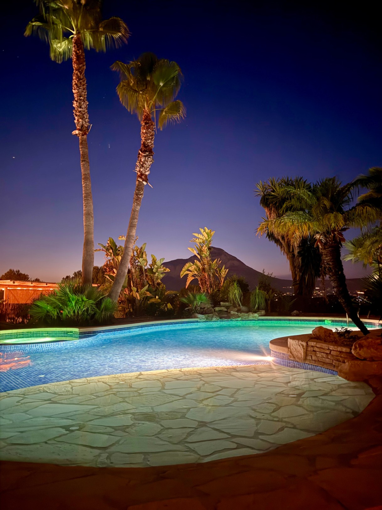 Piscina iluminada al anochecer con vistas directas a la silueta del Montgó, rodeada de vegetación tropical y pavimentos de piedra natural.