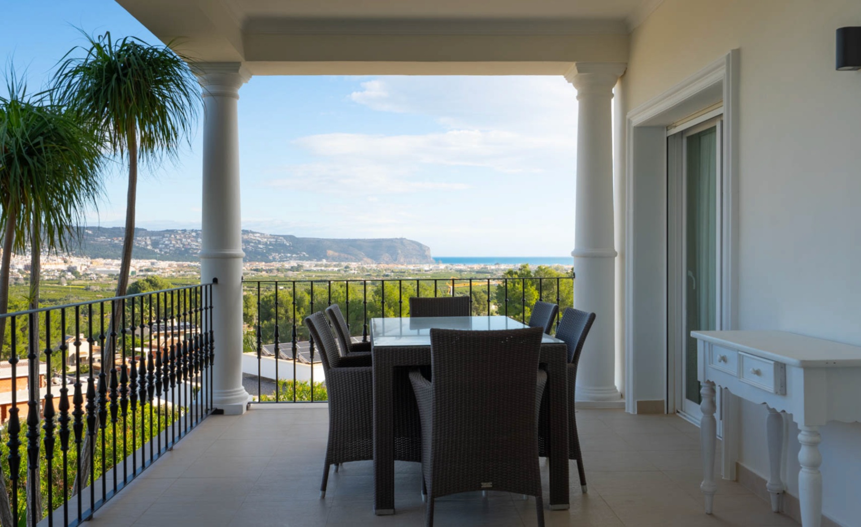 Terraza cubierta con columnas clásicas y barandilla de forja, equipada con comedor exterior y vistas panorámicas al mar y al valle de Jávea.