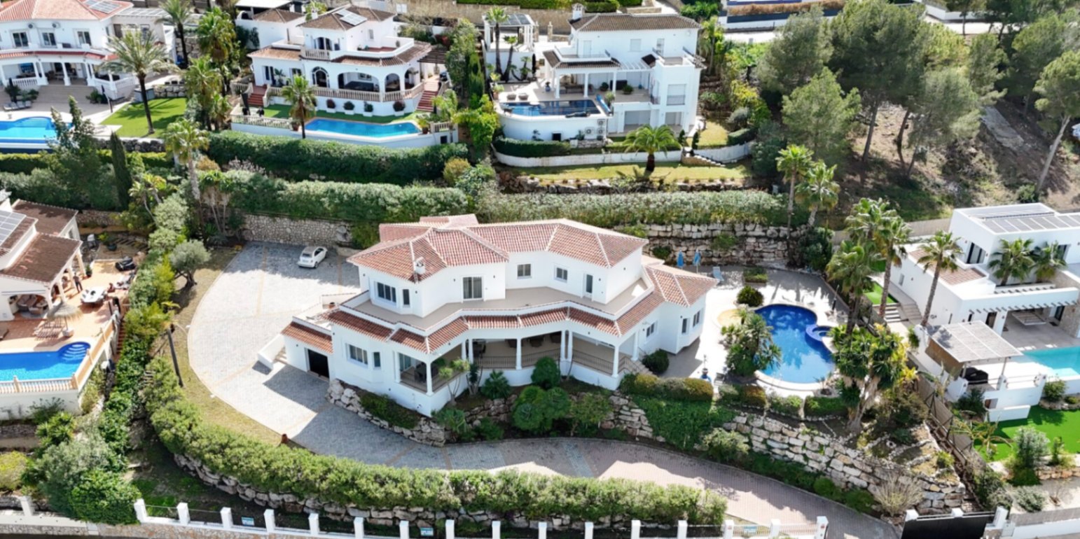 Vista aérea de la villa con tejados de teja, amplia entrada pavimentada, piscina privada rodeada de palmeras y terrazas en varios niveles sobre muros de piedra natural.