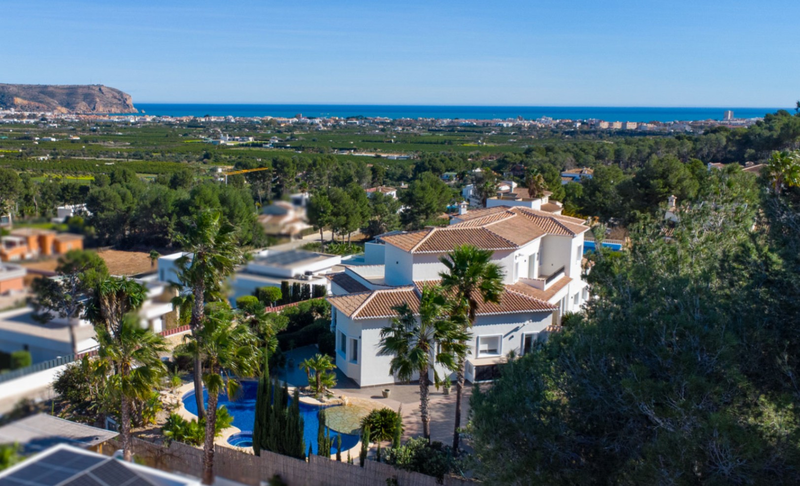 Vista aérea de la villa con tejados de teja cerámica, piscina de diseño orgánico y vistas panorámicas al mar Mediterráneo y la costa de Jávea.