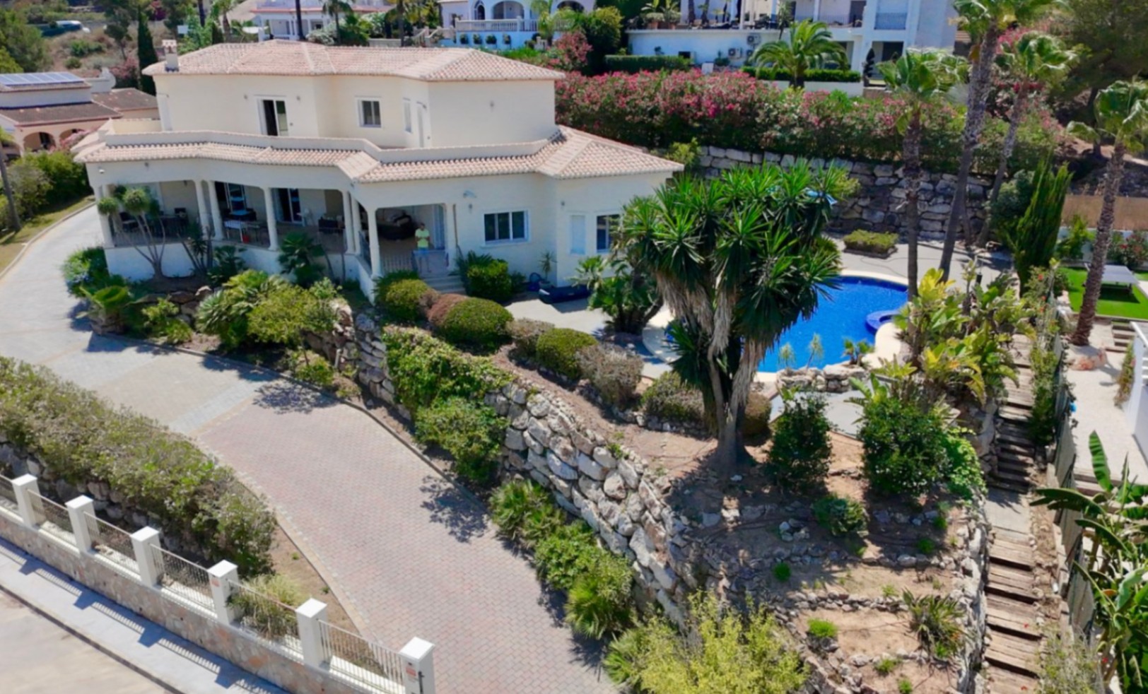 Vista exterior de la villa con fachada blanca y tejados de teja, destacando el acceso pavimentado, muros de piedra natural y jardines con palmeras y piscina.