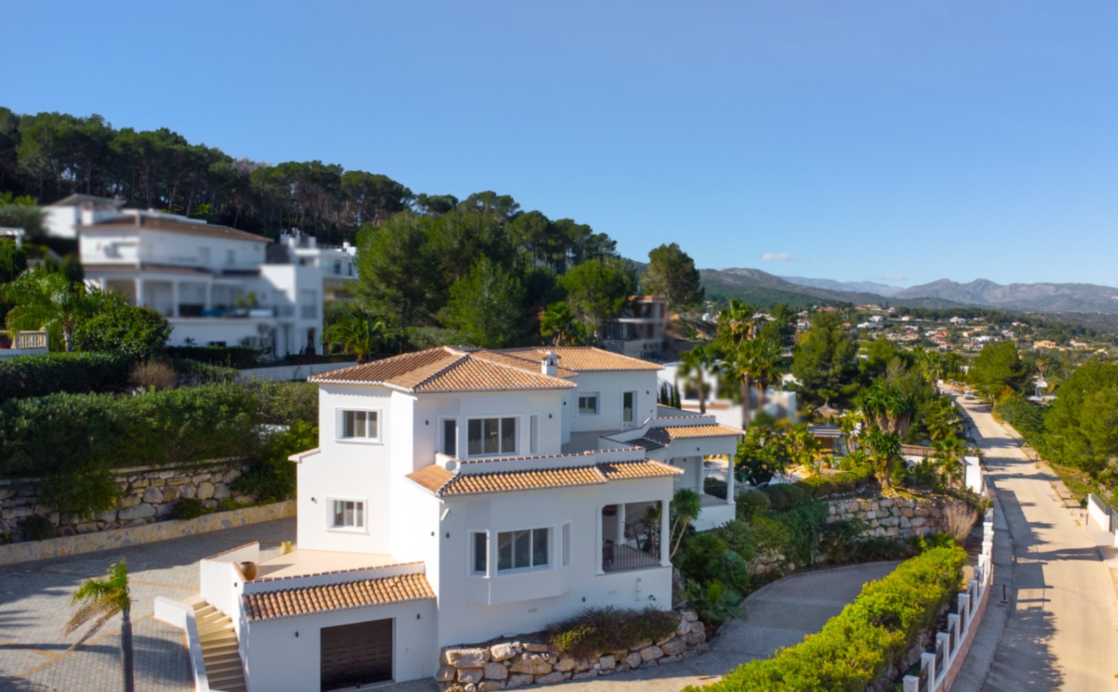 Vista exterior de la villa mediterránea con fachada blanca, tejados de teja cerámica y muros de piedra natural, situada en una parcela elevada con vistas a las montañas.